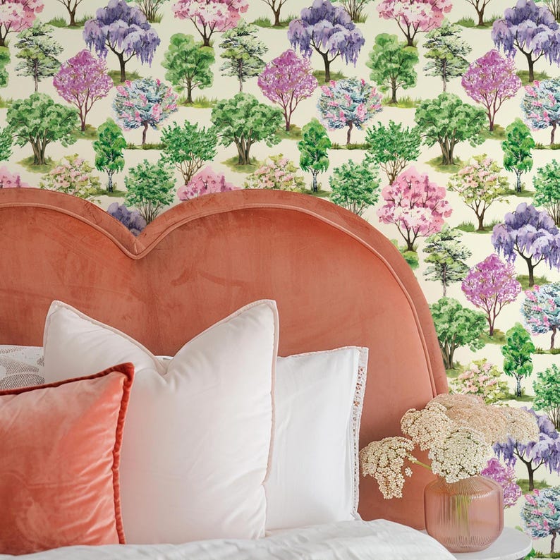 May include: A bedroom scene featuring a coral-colored, heart-shaped headboard, white pillows, and a coral-colored pillow. The wallpaper showcases a pattern of colorful trees in shades of green, purple, and pink. A vase of flowers sits on a nightstand.