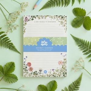 May include: A notepad with the text "A Few Things to Remember" and "Secret Garden" surrounded by floral illustrations. The notepad has lined pages and is decorated with a blue and white scalloped border. Green leaves and a pencil are scattered around the notepad.