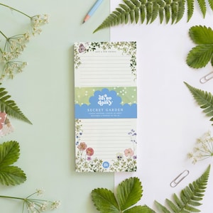 May include: A lined notepad with the text "Secret Garden" and "Oh So Daisy" in a floral design. The notepad is surrounded by green leaves, white flowers, a pencil, and paper clips.