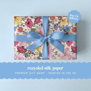 Puede incluir: Una caja de regalo rectangular envuelta en papel floral, atada con una cinta azul claro. El papel presenta un estampado de flores rosas, amarillas y azules sobre un fondo crema. El texto en la imagen dice "recycled silk paper" y "premium gift wrap - printed in the UK."