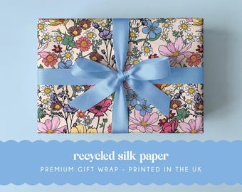 May include: A rectangular gift box wrapped in light pink paper featuring a colorful floral pattern of daisies, cosmos, and wildflowers, tied with a smooth light blue satin ribbon bow. The text reads recycled silk paper PREMIUM GIFT WRAP - PRINTED IN THE UK.