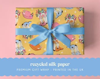 May include: A rectangular gift box wrapped in yellow paper featuring a repeating pattern of pink cowboy hats and white cowboy boots filled with colorful flowers, tied with a light blue satin ribbon. The text reads recycled silk paper.