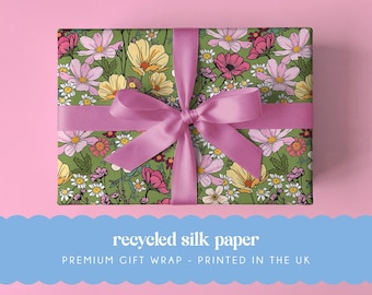 May include: A rectangular gift box wrapped in green paper featuring a colorful floral pattern of pink, yellow, and white flowers. A wide, smooth, light pink ribbon is tied in a bow around the center of the box. The text reads recycled silk paper.