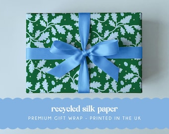 May include: A rectangular gift wrapped in dark green paper featuring a repeating pattern of light blue oak leaves and small yellow stars, tied with a wide light blue satin ribbon bow. The text reads recycled silk paper PREMIUM GIFT WRAP - PRINTED IN THE UK.