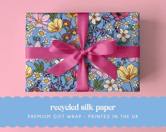 May include: A rectangular gift box wrapped in light blue paper featuring a colorful floral pattern of daisies, cosmos, and wildflowers. A vibrant magenta satin ribbon is tied in a bow around the center of the box. The text reads recycled silk paper.
