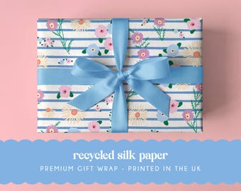 May include: A rectangular gift box wrapped in white paper with horizontal blue stripes and a floral pattern of pink, purple, and yellow flowers. A light blue satin ribbon is tied in a bow around the center. Text reads recycled silk paper.