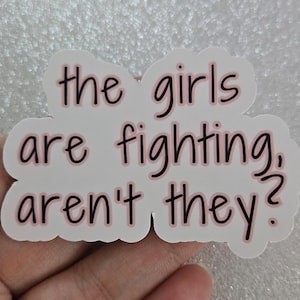 May include: A white sticker with a pink border and the phrase "the girls are fighting, aren't they?" in black and pink lettering. The sticker is in a hand.