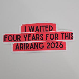 May include: A sticker with the text "I WAITED FOUR YEARS FOR THIS ARIRANG 2026" in black letters on a red background. The sticker has a white border and is on a white surface.