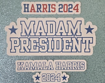 May include: A beige sticker with the text "HARRIS 2024" in red and blue lettering. Below the text is a larger beige sticker with the text "MADAM PRESIDENT" in blue lettering with a star on each side. Below that is a smaller beige sticker with the text "KAMALA HARRIS 2024" in blue lettering with a star on each side.