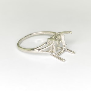 May include: A silver ring setting with a four-prong design. The ring features a delicate band that splits into a double-band design on each side of the setting. The setting is designed to hold a square or rectangular gemstone.