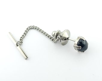 May include: A silver-toned tie tack with a black, rounded stone. The tie tack has a chain connecting the decorative head to a bar-shaped clasp. The stone is held in place by a silver metal setting.