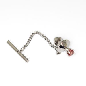 May include: A silver tie pin with a small, round, pink gemstone. The pin has a chain connecting the decorative head to a T-bar clasp. The background is plain white.