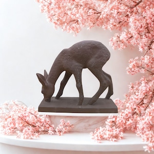 Könnte beinhalten: Eine bronzefarbene Hirschskulptur in einer anmutigen Pose, den Kopf gesenkt, als würde er trinken. Der Hirsch steht auf einem rechteckigen Sockel. Der Hintergrund zeigt rosa Kirschblüten.