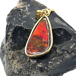 Könnte beinhalten: Ein tropfenförmiger Anhänger mit einem mehrfarbigen Edelstein in einem goldfarbenen Rahmen. Der Edelstein zeigt Farbtöne von Rot, Orange und Braun. Der Anhänger hängt an einer zarten goldfarbenen Kette, ein einzigartiges Schmuckstück.