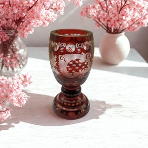 Könnte beinhalten: Ein tiefroter Glaskelch mit komplexen Ätzmustern, darunter florale Motive, Gitterdesigns und kreisförmige Akzente. Das Glas besitzt einen dicken, abgerundeten Stiel und einen breiten, stabilen Fuß, was den Kontrast zwischen dem dunkelroten Glas und den weißen Ätzdetails hervorhebt.