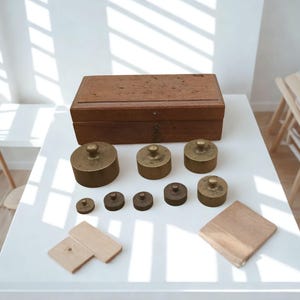May include: An assortment of antique brass weights of different sizes, displayed on a white surface. A wooden box and small wooden blocks are also visible. The weights are likely for measuring mass.