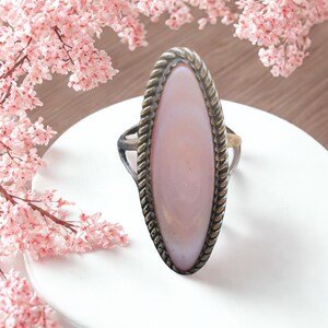Peut inclure: Bague ovale avec une pierre rose pâle, sertie dans un cadre en métal argenté. Le cadre présente un motif en forme de corde autour de la pierre. La bague est posée sur une surface blanche, avec des fleurs roses en arrière-plan.
