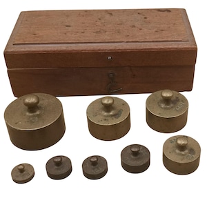 Könnte beinhalten: Ein antikes Set aus Messinggewichten mit einer Holzkiste. Die Gewichte sind zylindrisch mit einem kleinen Knopf oben. Die Kiste ist rechteckig und aus Holz. Die Gewichte sind unterschiedlich groß und vor der Kiste angeordnet.