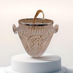 Könnte beinhalten: Ein klarer Eisbehälter aus Glas mit silbernem Rand und goldfarbenem Griff. Der Eimer hat ein Rautenmuster und zwei seitliche Griffe. Der Eisbehälter wird auf einem weißen Sockel präsentiert.