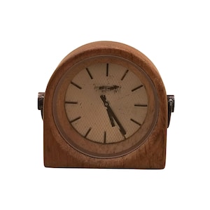 May include: A tabletop clock with a rounded wooden frame and a light-coloured, textured face. The clock features thin, dark hour markers, dark hands, and a metallic hinge detail on each side. The face shows signs of wear with visible markings.