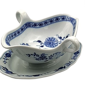 Puede incluir: Salsera y platillo de porcelana blanca con un diseño floral y de vid azul. La salsera tiene un asa curva a cada lado. Los bordes de la salsera y el platillo son festoneados.