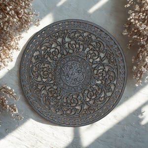 May include: A circular decorative metal plate with an elaborate openwork design. The plate has a central floral pattern, surrounded by swirling motifs and a detailed border. The colour is a muted grey, with subtle tonal variations, and it rests on a white surface.