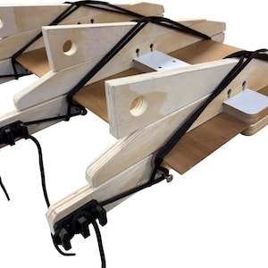 May include: A wooden structure with multiple light-colored, rectangular pieces connected by black cords. The pieces have circular holes and are attached to black plastic components. The structure appears to be a woodworking tool or jig.