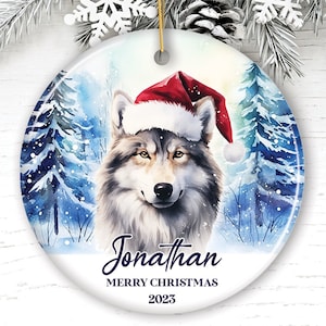 May include: A white ceramic Christmas ornament with a watercolor painting of a wolf wearing a Santa hat. The wolf is surrounded by blue and white trees. The text "Jonathan Merry Christmas 2023" is printed on the ornament.