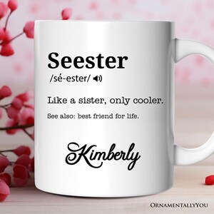 以下が含まれることがあります： 白いセラミック製のマグカップに、黒い文字で「Seester /sé-ester/ 姉妹のような存在、でももっとクール。参考：生涯の親友。Kimberly」と書いてあります。