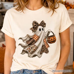 May include: A cream-coloured t-shirt featuring a graphic of a ghost wearing a leopard print bow. The ghost is depicted with patchwork patterns in orange, brown, and white, and is holding a small pumpkin-shaped trick-or-treat bucket.