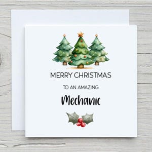 May include: A white Christmas card with the message "MERRY CHRISTMAS TO AN AMAZING Mechanic." The card displays three green Christmas trees with gold star decorations and baubles. A sprig of holly with red berries is at the bottom.