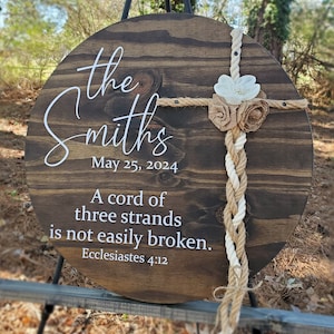 May include: A round wooden sign with the text "the Smiths May 25, 2024 A cord of three strands is not easily broken. Ecclesiastes 4:12" and a white and brown rope with a white flower and brown burlap flowers.