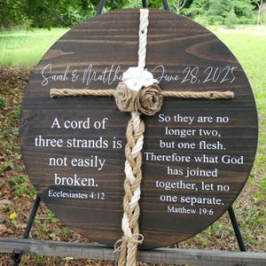 May include: A round, dark stained wooden sign with a cross design made of rope and fabric flowers. The sign includes the names "Sarah & Matthew" and the date "June 28, 2025". It also features Bible verses about marriage.