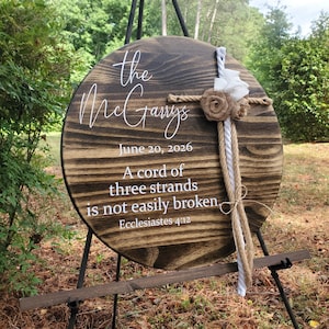 May include: A round, dark-stained wooden sign featuring white text that reads: The McGarrys, June 20, 2026, A cord of three strands is not easily broken, Ecclesiastes 4:12. A decorative cross made of white and jute rope with burlap flowers is attached.