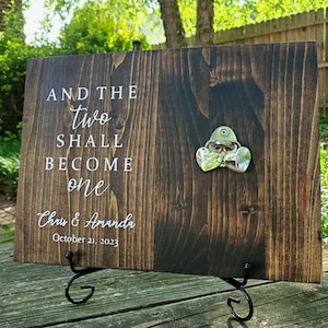 May include: A dark stained wooden sign with the text "AND THE TWO SHALL BECOME ONE" and the names "Chris & Amanda" with the date "October 21, 2023". A metal bottle opener is attached. The sign is displayed on a black metal stand.