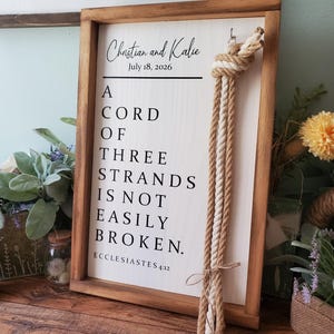 May include: A rectangular wooden sign with a light-colored face and dark wood frame. The text reads: Christian and Kalie, July 18, 2026, A CORD OF THREE STRANDS IS NOT EASILY BROKEN. ECCLESIASTES 4:12. Three braided ropes are knotted and hung on the right side.