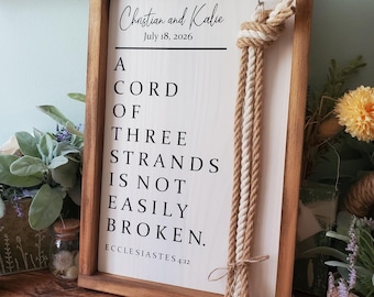 May include: A rectangular wooden sign with a light-colored face and dark wood frame. The text reads: Christian and Kalie, July 18, 2026, A CORD OF THREE STRANDS IS NOT EASILY BROKEN. ECCLESIASTES 4:12. Three braided ropes are knotted and hung on the right side.