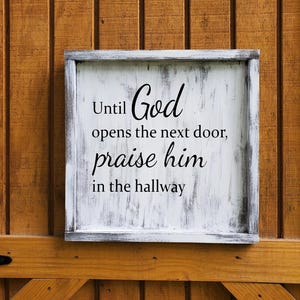 May include: A square, white-painted wooden sign with a gray frame. The sign displays the text "Until God opens the next door, praise him in the hallway" in black script. The sign is mounted on a wooden door.