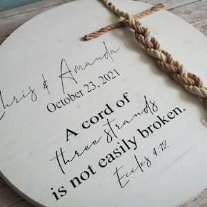 May include: White wooden round sign with the text "Chris & Amanda" and the date "October 23, 2021". The sign also includes the quote "A cord of three strands is not easily broken. Eccles 4:12" and a braided rope.
