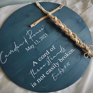 May include: A round wooden sign with a teal blue background and white lettering. The sign says "Camden & Rence May 13, 2023" and "A cord of three strands is not easily broken. Eccles 4:12". A braided rope is attached to the top of the sign.