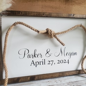 May include: A white wooden sign with a brown frame and a rope tied in a knot at the top. The sign says "Parker & Megan April 27, 2024".