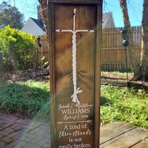 May include: A wooden sign with a white rope tied in a cross shape. The sign has the text "Sarah & Matthew WILLIAMS April 6, 2024 A cord of three strands is not easily broken. Ecclesiastes 4:12"