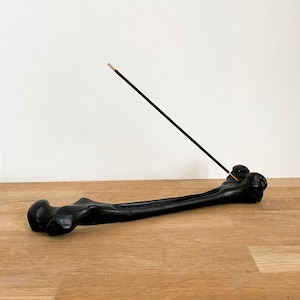 以下が含まれることがあります： 黒い骨の形をしたお香立てに、黒いお香が燃えています。お香立ては木製の表面に置かれています。