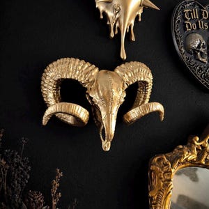 May include: A gold-coloured ram skull wall decoration with large, curved horns. The skull is mounted on a black wall, alongside other gold and black gothic-themed decorations. The skull is a detailed, textured sculpture.