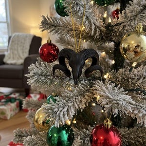 Peut inclure: Ornement de Noël en forme de crâne de bélier noir suspendu à un sapin enneigé, entouré de boules rouges, vertes et dorées. L'arbre est décoré de flocons de neige blancs et de lumières, créant une scène de fête.