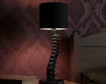 May include: A unique black table lamp with a spine-shaped base and a black lampshade. The lamp is on a wooden nightstand next to a book. The background features a patterned burgundy wallpaper.