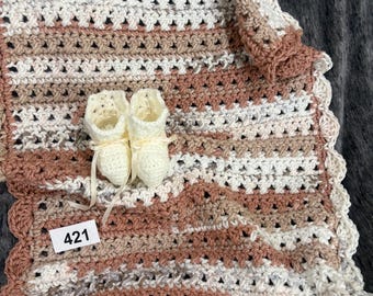 May include: A handmade crochet baby blanket featuring horizontal stripes in shades of cream, tan, and dusty rose with a scalloped edge. Two matching cream-colored crochet baby booties with ribbon ties are placed on top. A small white tag displays the number 421.