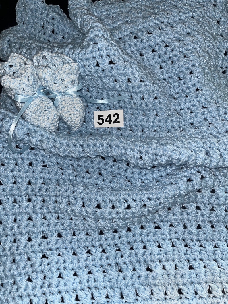 May include: A light blue crocheted baby blanket with matching booties. The blanket has a simple, repeating pattern. The booties have a white and blue color scheme. The tag on the blanket reads "542".