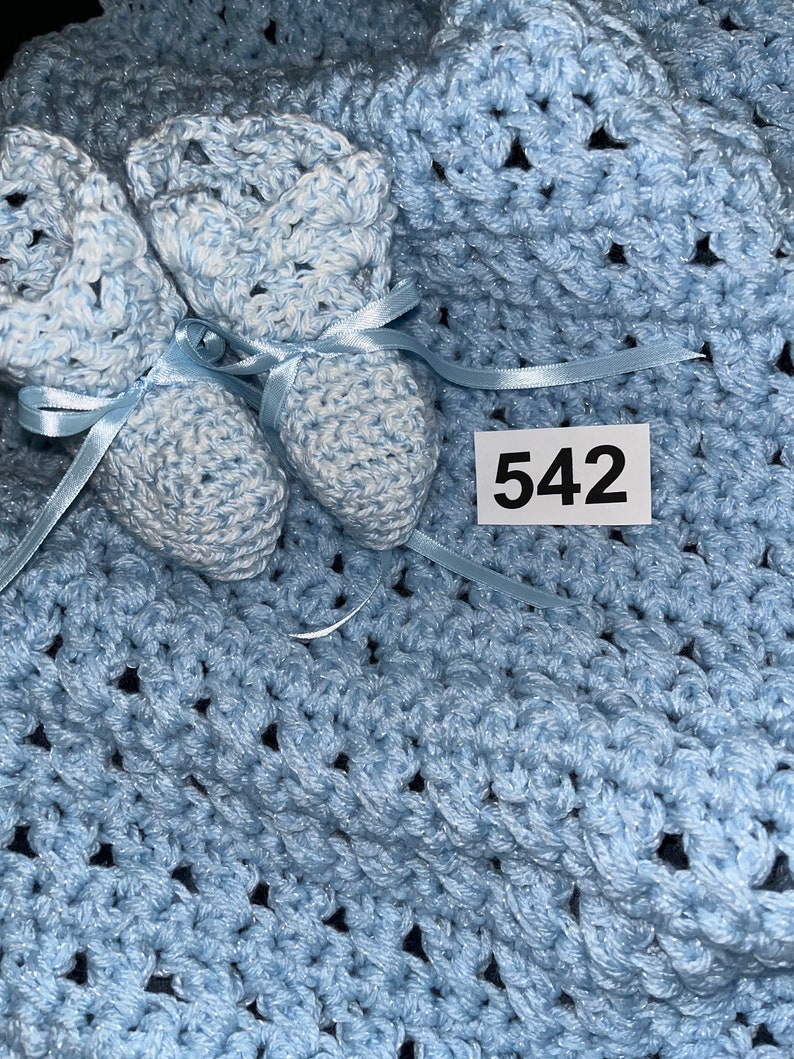 May include: A light blue crocheted baby blanket with two matching baby booties. The blanket has a textured pattern and the booties have white accents. The number 542 is written on a white tag.