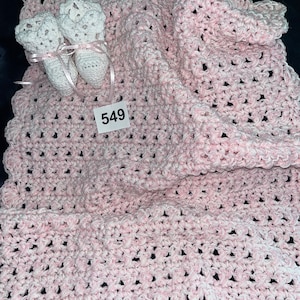 May include: A pink crocheted baby blanket with a scalloped edge and two white crocheted booties. The blanket has a tag with the number 549 on it.